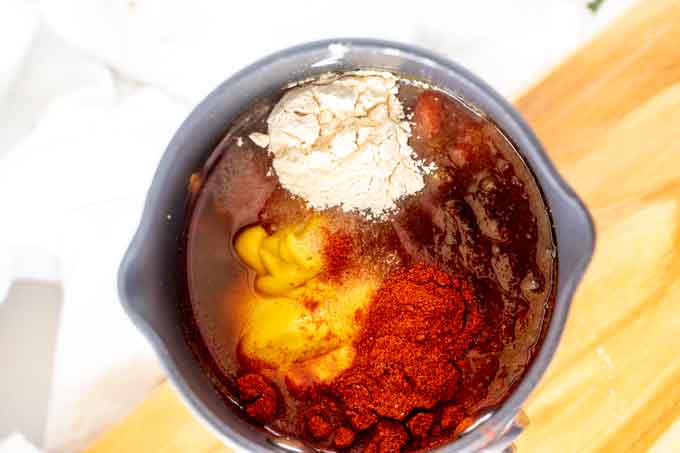 Photo of ketchup, yellow mustard, apple cider vinegar, brown sugar substitute, smoked paprika, liquid smoke, garlic powder, and salt in a saucepan.