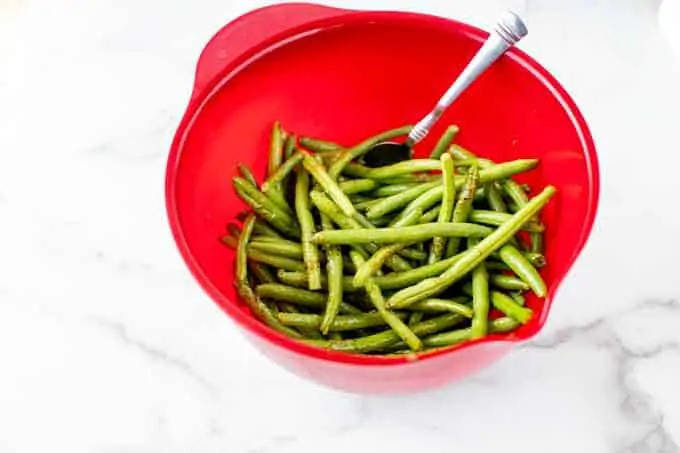 Photo of a red bowl with green beans that have been tossed with oil and seasonings.