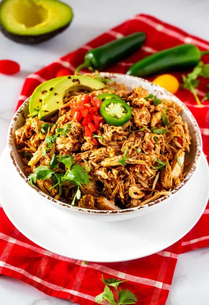 White bowl of slow cooker chicken tinga garnished with avocado, jalapeno, tomato, and cilantro sitting on a red napkin. 