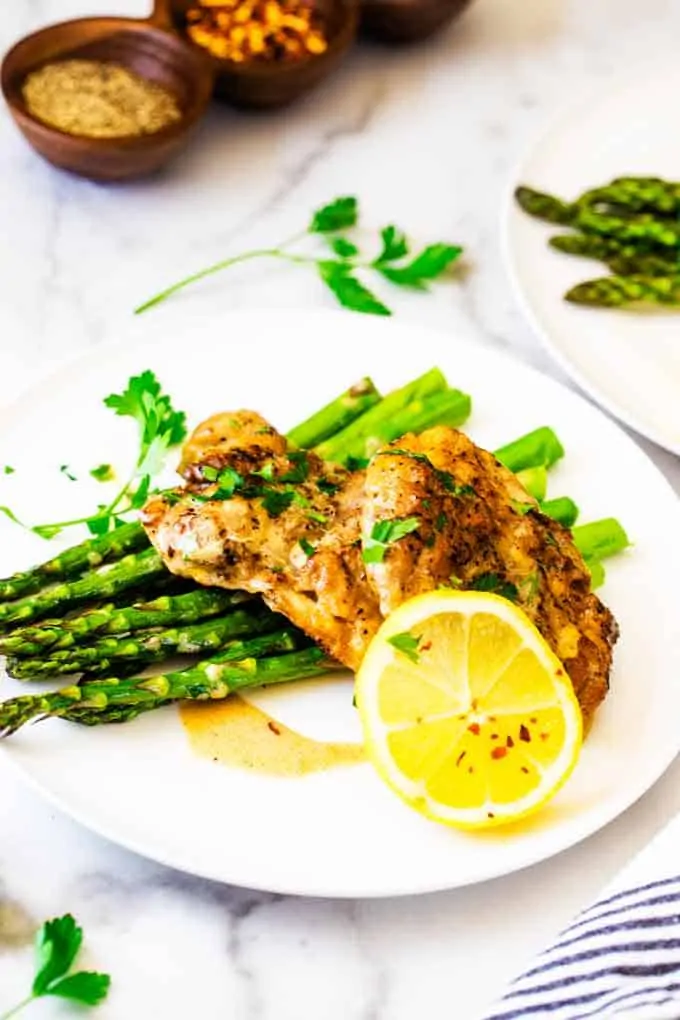 Photo of Instant Pot Lemon Chicken on a white plate with asparagus garnished with parsley, crushed red pepper flakes, and a lemon.