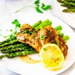Square photo of Instant Pot Lemon Chicken on a white plate with asparagus garnished with parsley, crushed red pepper flakes, and a lemon.