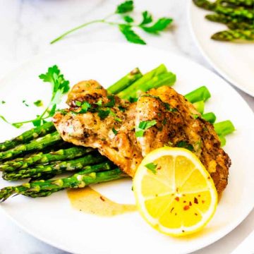 Square photo of Instant Pot Lemon Chicken on a white plate with asparagus garnished with parsley, crushed red pepper flakes, and a lemon.