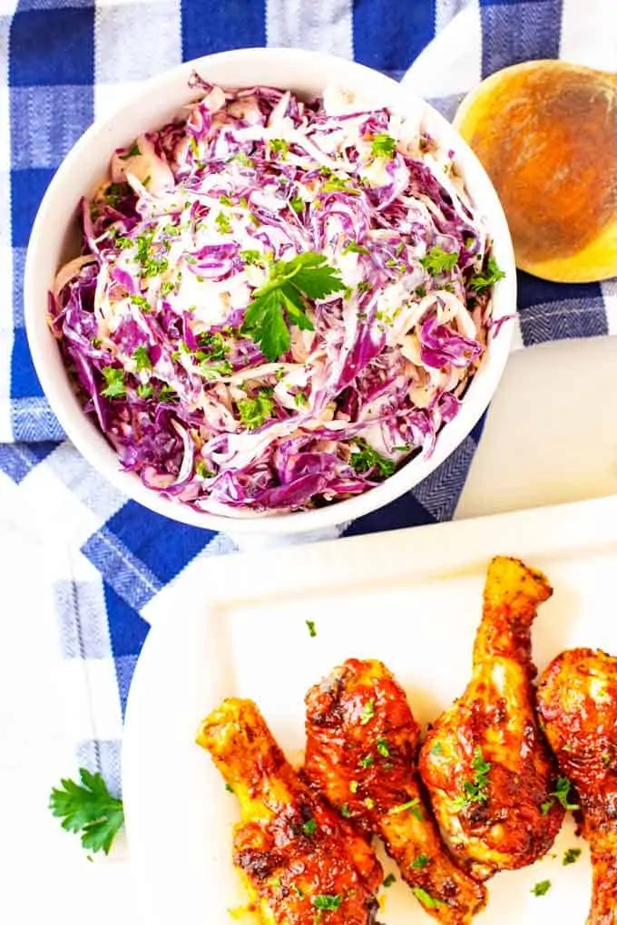 Overhead photo of a white bowl of keto coleslaw sitting on a blue and white checked napkin with a platter of BBQ Chicken next to it.