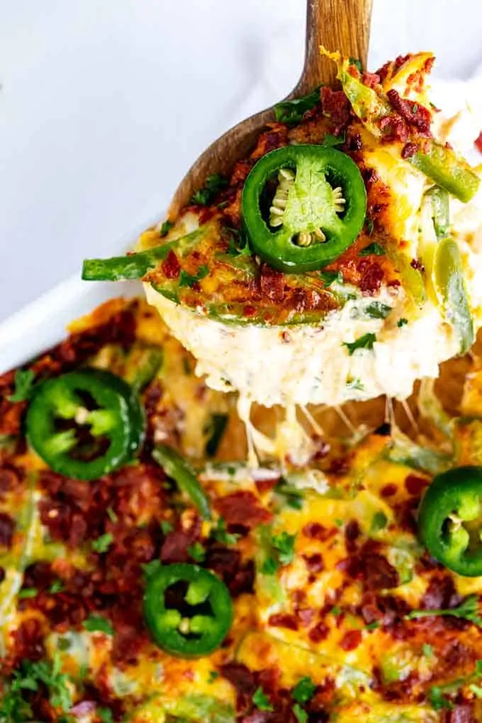 Photo of Jalapeno popper casserole in a white casserole dish garnished with fresh jalapenos with a spoonful being taken out of it with a cheese pull.