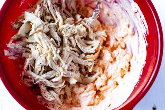 Photo of shredded chicken being mixed into a creamy sauce in a red bowl.