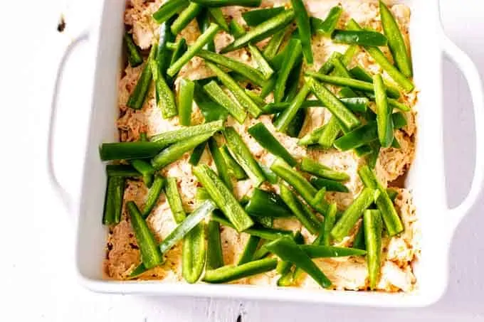 Photo of sliced jalapenos on top of a creamy chicken mixture in a white casserole dish.