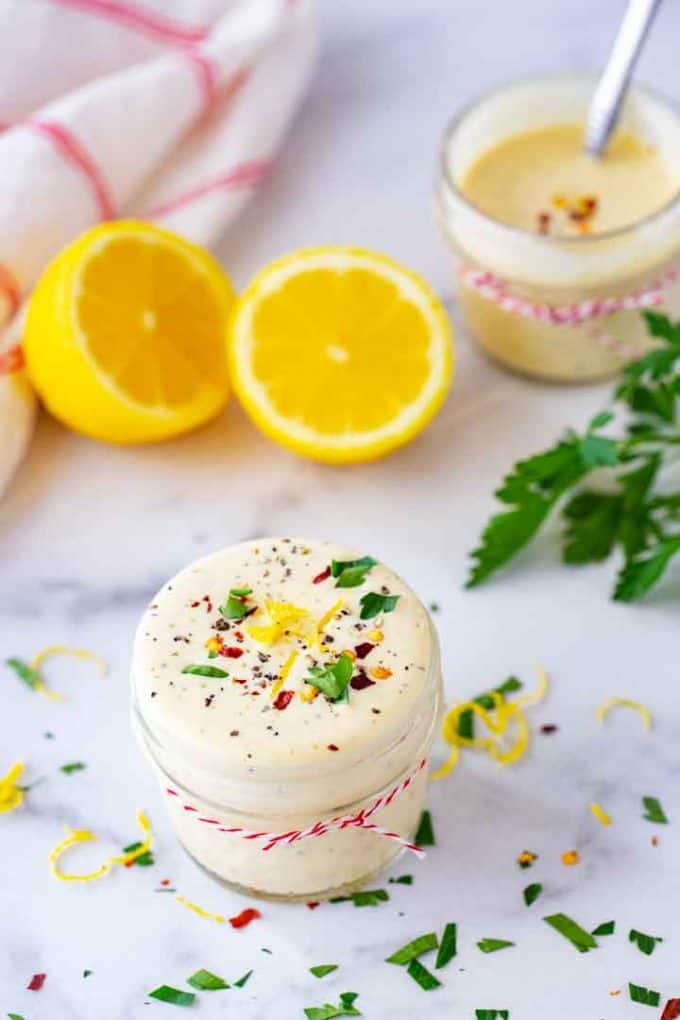 Photo of lemon aioli on a white background with lemon zest, parsley, and red pepper flakes as a garnish and a halved lemon and another jar of aioli with a spoon in the background.