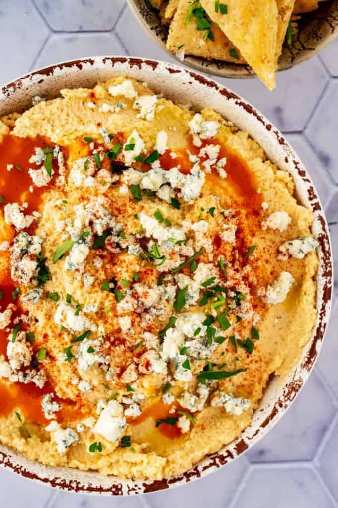 Close up overhead photo of a rustic white bowl with cauliflower hummus in it garnished with buffalo sauce, blue cheese, and parsley.