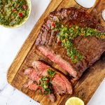 Square overhead photo of keto flank steak with three thin slices cut from it on a wooden cutting board garnished with chimichurri.