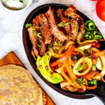 Square overhead photo of a skillet of keto steak fajitas with tortillas sitting next to it.