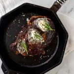 Square overhead photo of a seared ribeye in a cast iron skillet garnished with herb butter and rosemary.