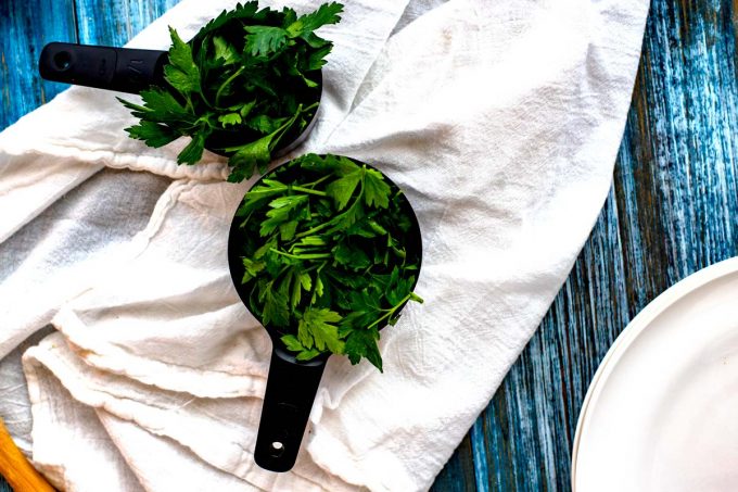Photo of 2 black measuring cups with parsley and cilantro in them on a white napkin.