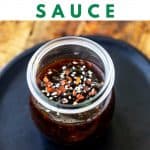 Photo of a small jar sitting on a black plate sprinkled with sesame seeds and crushed red pepper flakes with the text Keto Teriyaki Sauce above.
