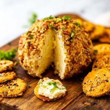 Square photo of a keto cheese ball with a slice taken out of it on a wooden cutting board surrounded by crackers.