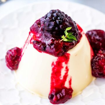 Square close up photo of a keto panna cotta drizzled in blackberry sauce on a white plate with a fork behind it.