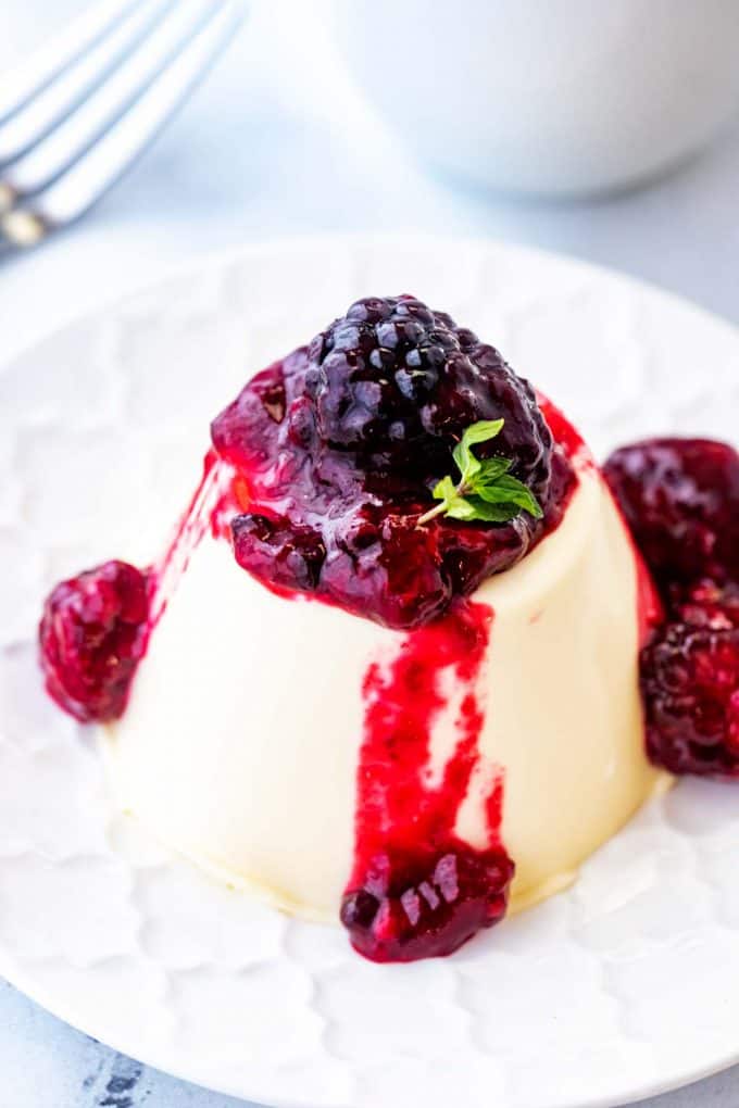 Close up photo of a keto panna cotta drizzled in blackberry sauce on a white plate with a fork behind it.