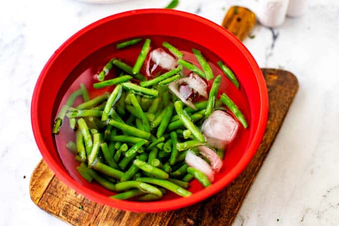 Photo of a red bowl with blanched green beans and ice cubes.