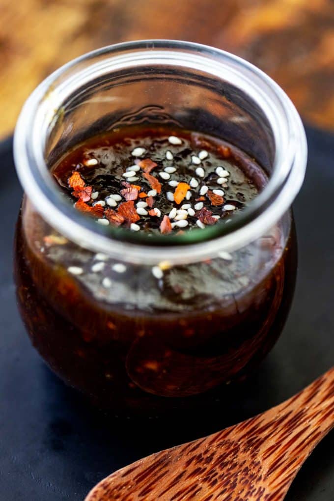 Close up photo of keto teriyaki sauce in a small glass jar garnished with red pepper flakes and sesame seeds with a wooden spoon sitting next to it.