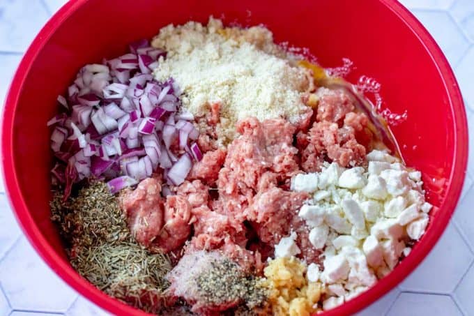 Photo of a red bowl with ground lamb, eggs, red onion, feta, almond flour, garlic, rosemary, oregano, salt, and pepper.