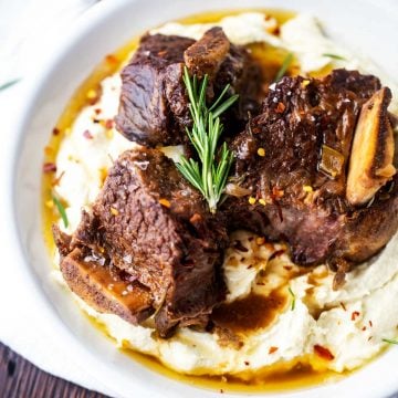 Square photo of a white plate of keto short ribs over mashed cauliflower garnished with rosemary.