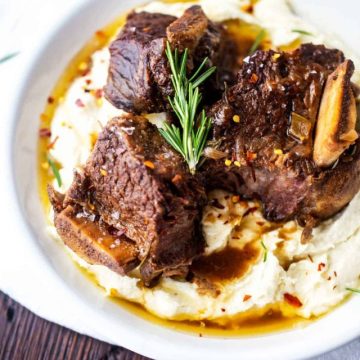 Photo of a white plate of keto short ribs over mashed cauliflower garnished with rosemary.