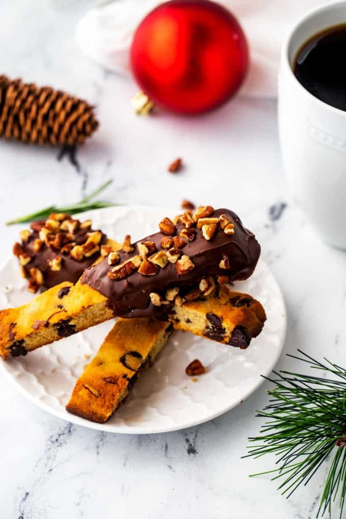Photo of keto biscotti on a white plate surrounded by a coffee cup, Christmas ornament, and pine cone.