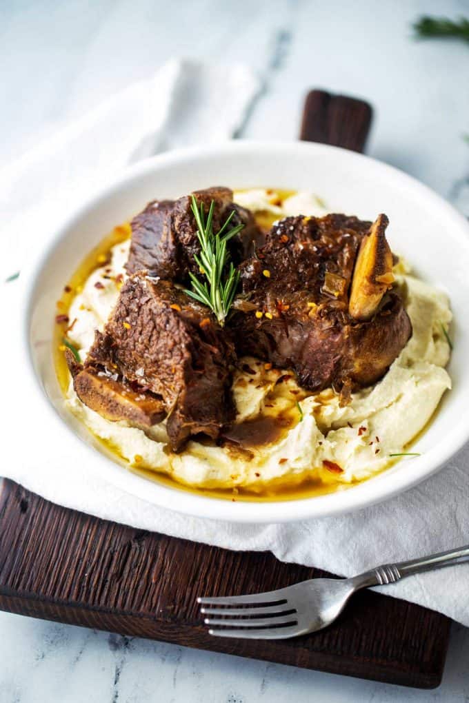 Photo of a white plate of keto short ribs over mashed cauliflower garnished with rosemary sitting on a wooden cutting board..