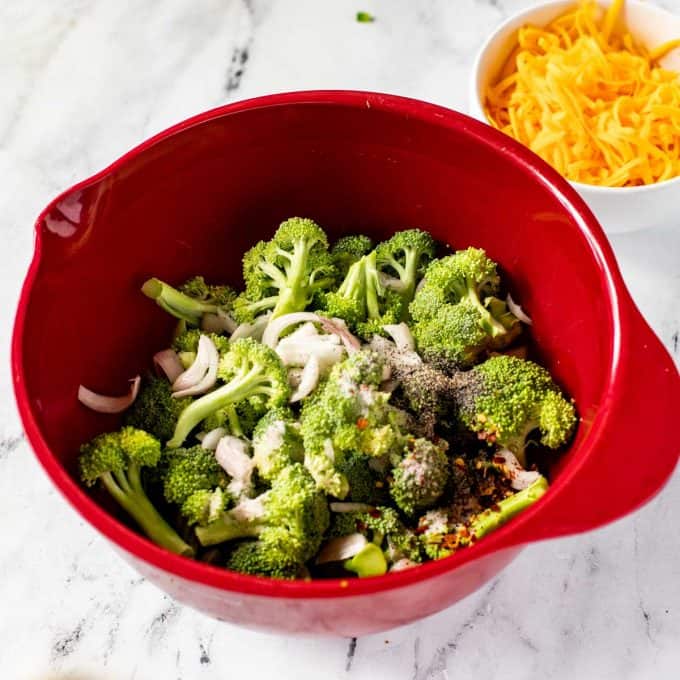 Red bowl of broccoli, seasonings, and shallots.