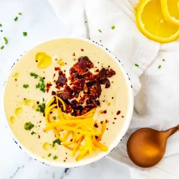 Square overhead photo of a white bowl with Keto Cauliflower Soup garnished with bacon, chives, and shredded cheddar cheese.