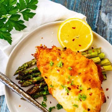 White plate with a piece of Keto Parmesan Crusted Chicken on a bed of asparagus.