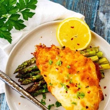 White plate with a piece of Parmesan Crusted Chicken on a bed of asparagus.