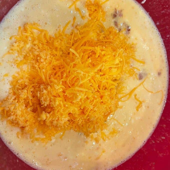 Photo of shredded cheddar cheese being added to a bowl of beaten eggs, heavy cream, sour cream, sausage, and seasonings.