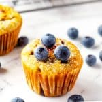 Square photo of a keto blueberry muffin topped with fresh blueberries and surrounded by more blueberries and muffins.