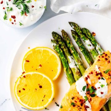 Photo of lemon dill sauce drizzled on salmon and asparagus with a small white bowl of sauce beside it.