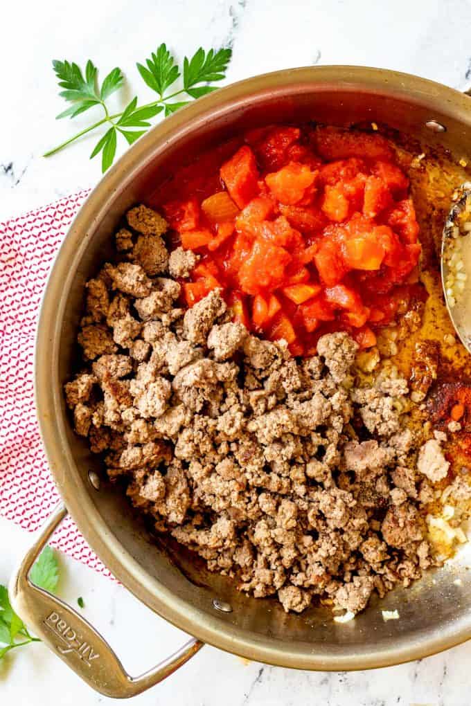 Photo of ground beef and chopped tomato in a large skillet with seasonings, onion, and garlic.