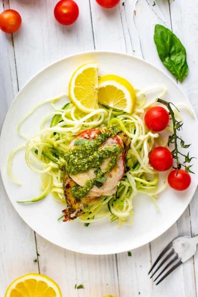 Photo of Keto Pesto Chicken sitting on a bed of zucchini noodles on a white plate with lemon and cherry tomatoes on the plate.