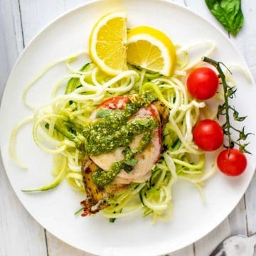 Square photo of Keto Pesto Chicken sitting on a bed of zucchini noodles on a white plate with lemon and cherry tomatoes on the plate.