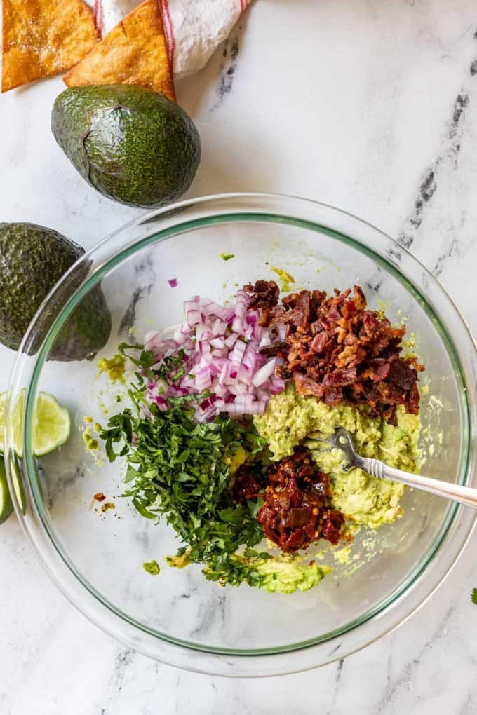 Photo of two mashed avocados being combined with lime juice, seasonings, bacon, onion, cilantro, and chipotle chiles in adobo sauce.