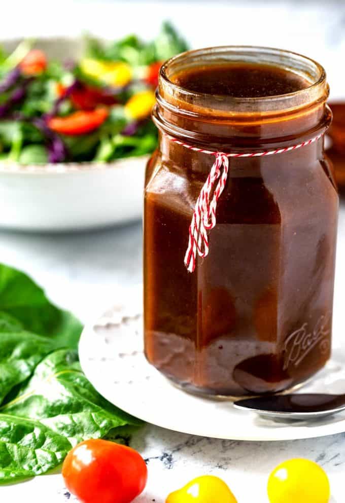 Close up photo of a jar of keto balsamic vinaigrette with tomatoes and greens close to it and a salad behind it.