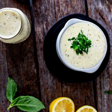 Square Close up overhead photo of Keto Alfredo Sauce in a small white crock on a black plate with a jar of Alfredo sauce next to it and basil, tomatoes and lemon surrounding it.