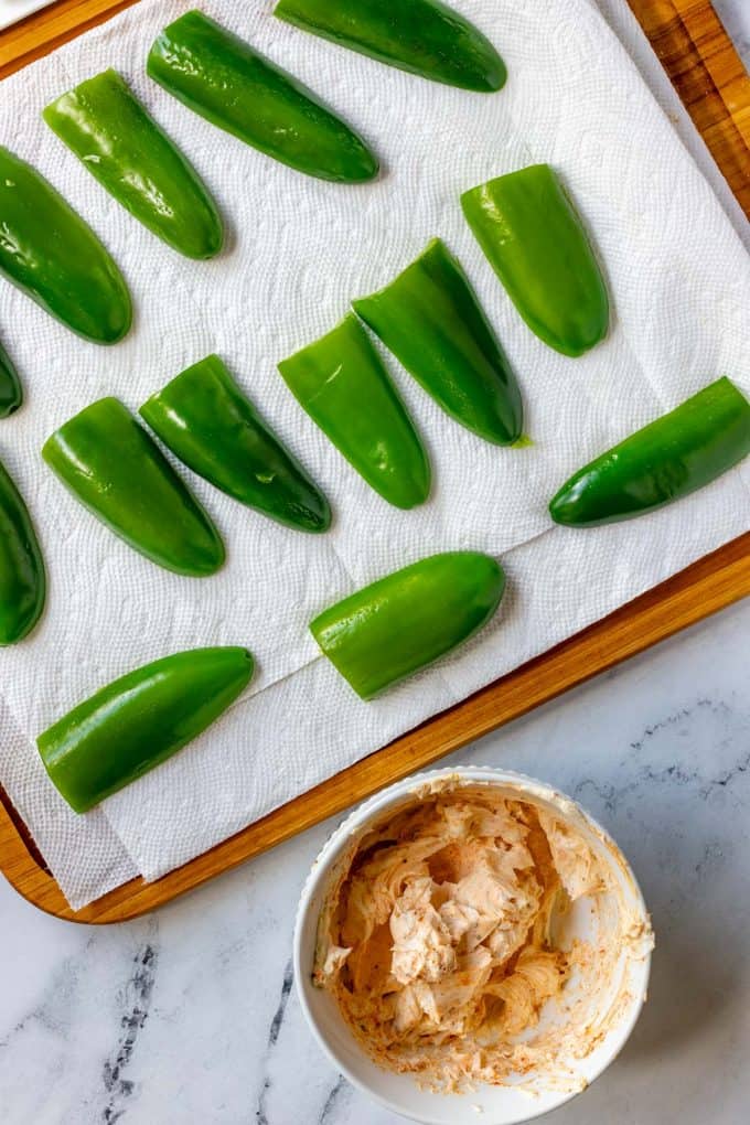 Jalapeno halves that have been partially roasted on paper towels with a cream cheese filling sitting next to them.
