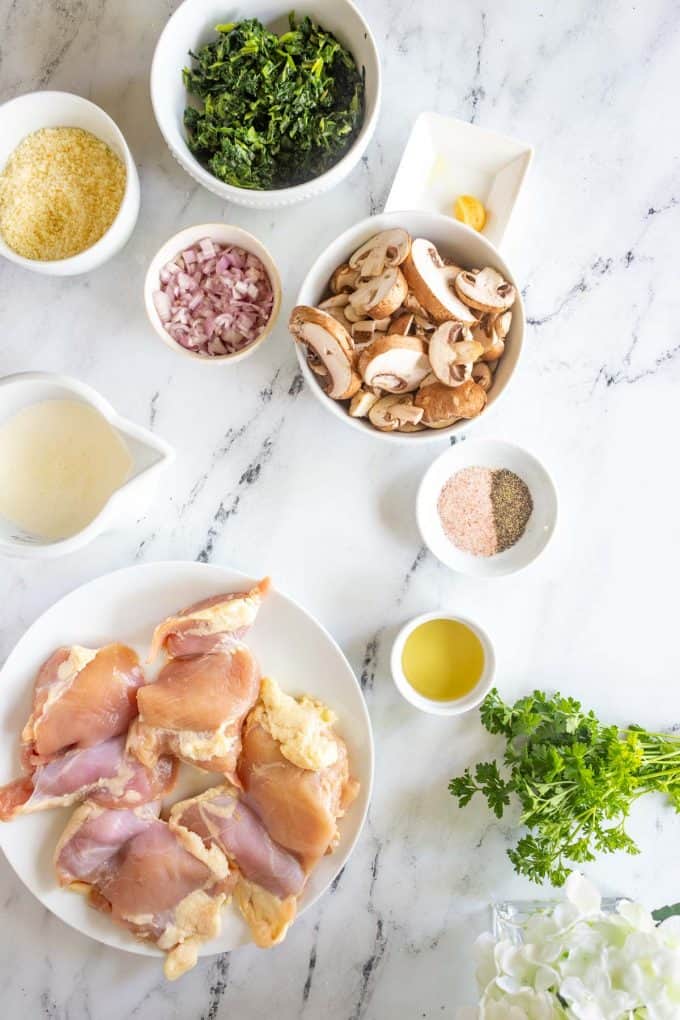 Overhead photo of chicken thighs, heavy cream, parmesan, shallot, spinach, garlic, mushrooms, seasonings, and avocado oil in prep bowls.