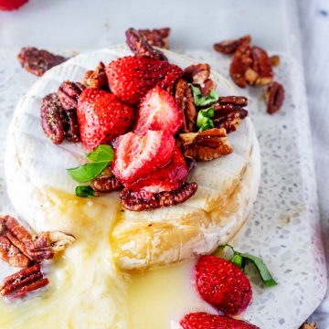 Square close up of a baked wheel of keto brie cheese with cheese oozing out of it topped with strawberries and pecans.