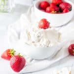 Square photo of keto whipped cream topped with a strawberry and a bowl of strawberries behind it.