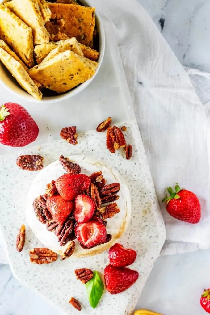 Overhead photo of keto brie topped with strawberries and pecans and a small bowl of crackers behind it.
