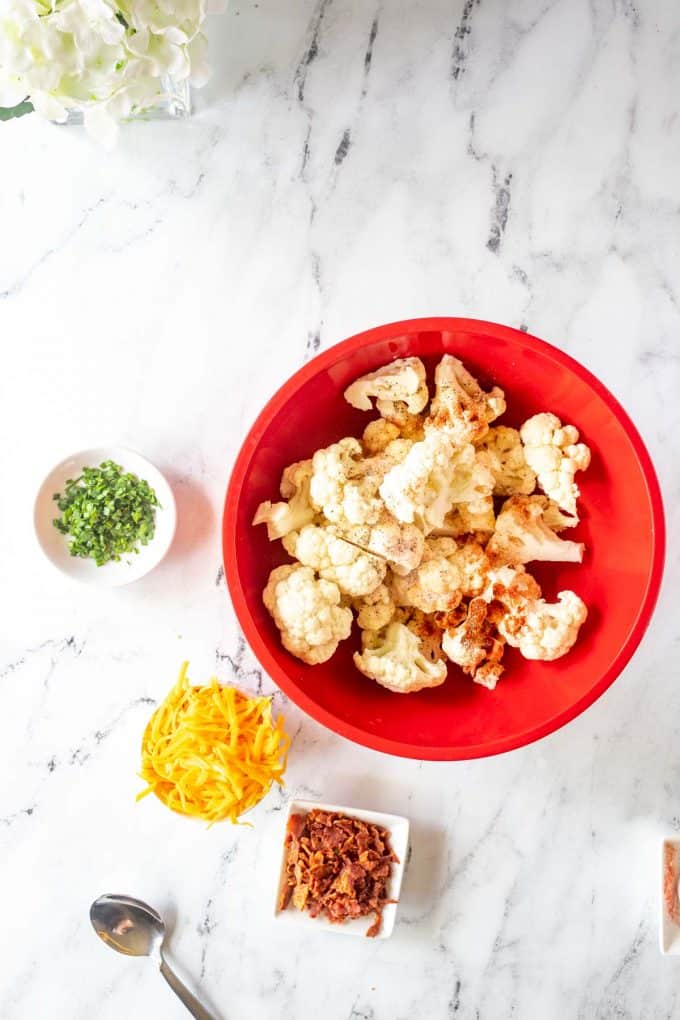 Overhead photo of a bowl of cauliflower being tossed with seasonings and oil and small prep bowls with chives, shredded cheese, and bacon sitting next to it.