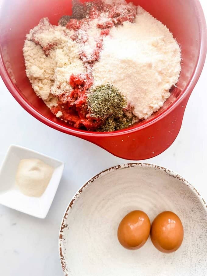 Overhead photo of a bowl with two eggs, a small dish of gelatin, and a large red bowl with ground beef, seasonings, parmesan, and almond flour.