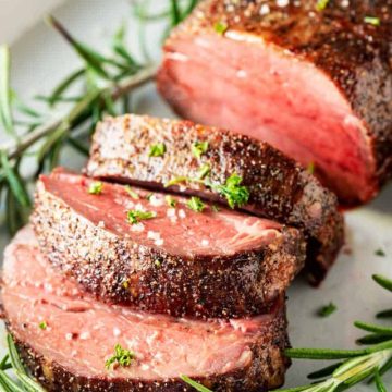 Square photo of a slow roasted beef tenderloin with three slices surrounded by rosemary on a platter.