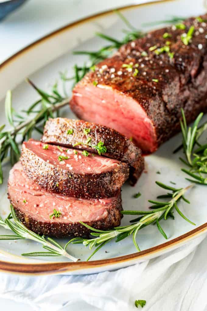 Photo of a slow roasted beef tenderloin with three slices surrounded by rosemary on a platter.