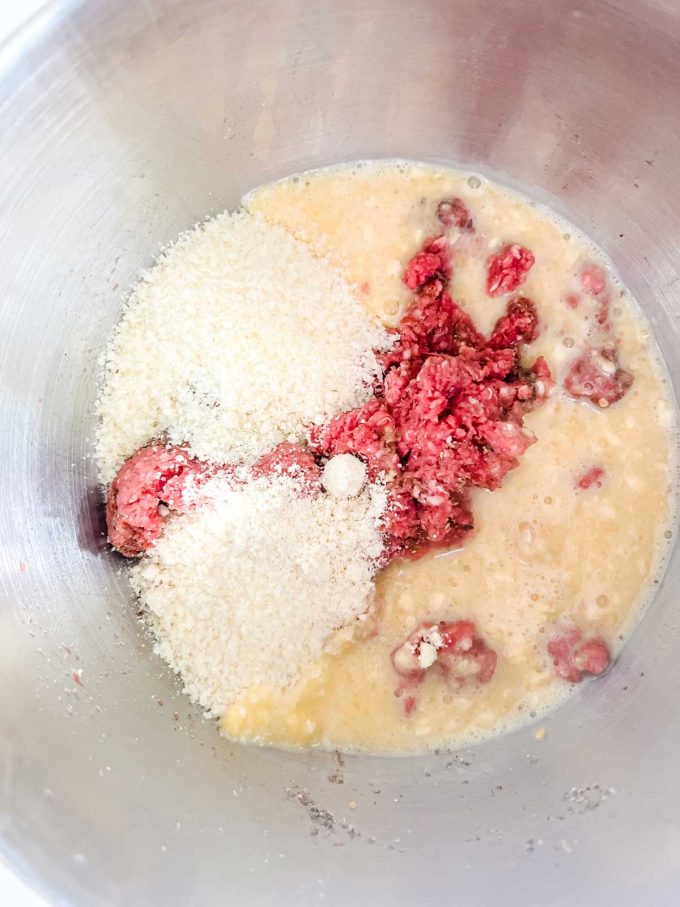 Photo of seasoning ground beef in a bowl that has had egg, gelatin, almond flour, and parmesan cheese sprinkled over it.